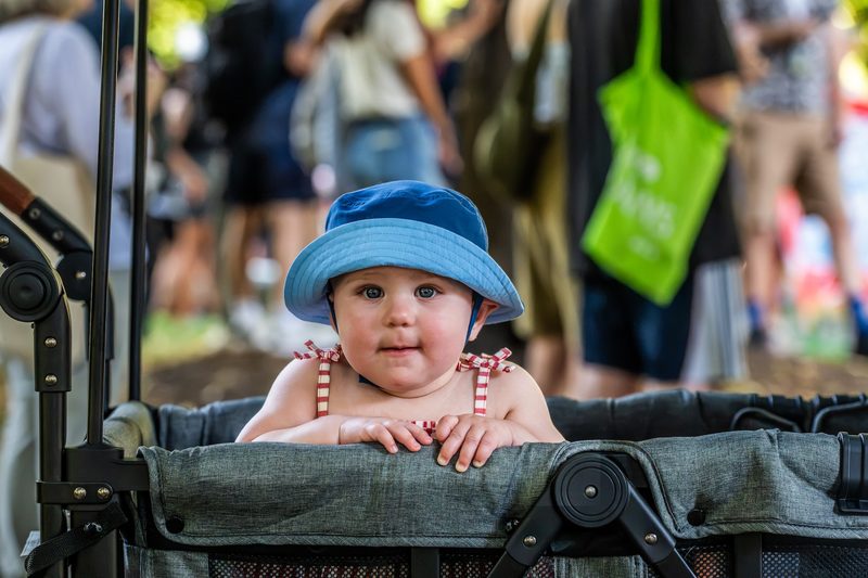 Baby in a blue sun hat sitting up in a stroller at a busy summer market
