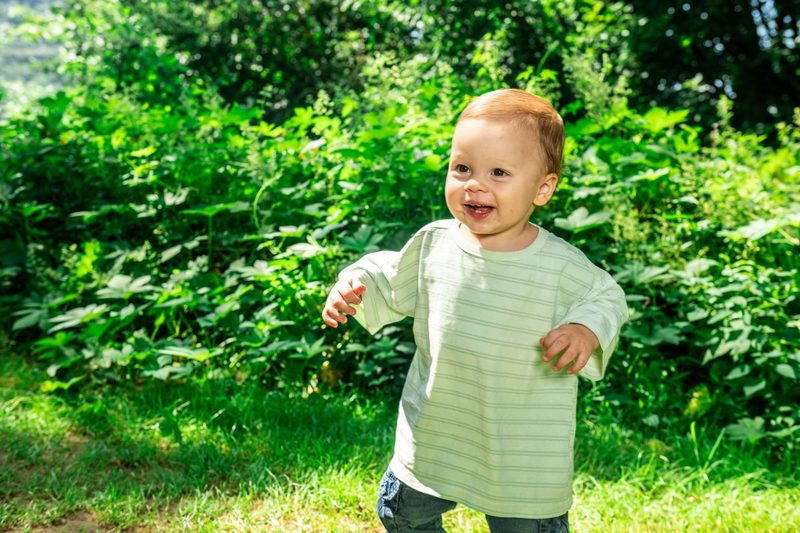 Baby in a striped shirt toddling through tall summer greenery, mid-laugh