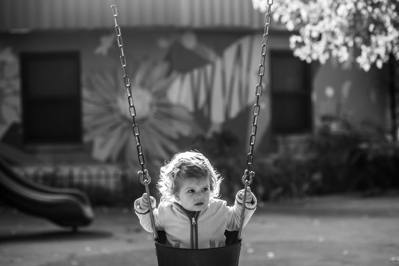 Black-and-white photograph of a baby sitting in a playground swing in front of a painted mural