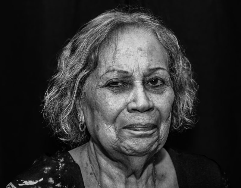 Black-and-white portrait of an older woman against a dark background, looking directly at the camera