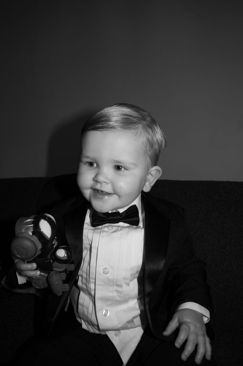 Black-and-white portrait of a small boy in a tuxedo and bow tie holding a toy camera