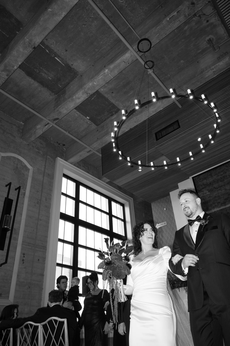 Couple walking arm in arm beneath a circular chandelier in a brick and concrete Philadelphia venue, photographed in black and white