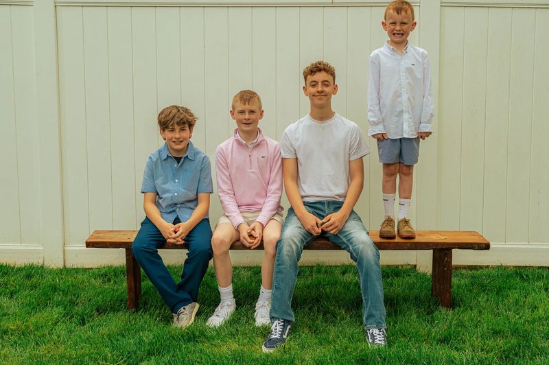 Four brothers on a wooden bench against a white fence, one standing and grinning, during a family photo session