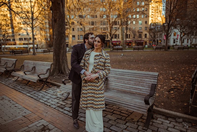 Engaged couple standing together beside a park bench in a Philadelphia square on an autumn afternoon