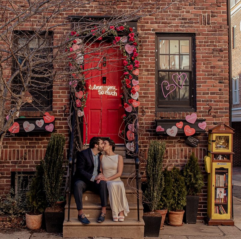 Couple kissing on a Philadelphia rowhouse stoop beside a red door covered in paper hearts