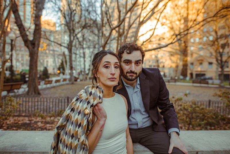 Close engagement portrait of a couple seated on a park bench with autumn trees behind them