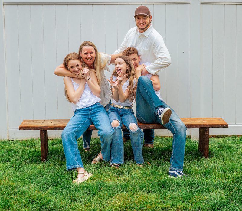 Family of five piled together on a bench against a white barn wall, laughing
