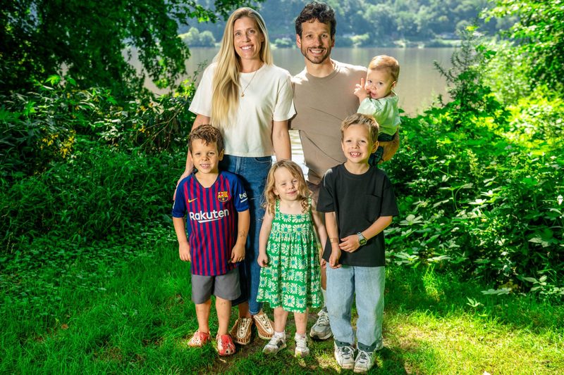 Family of six smiling together at the water's edge in summer, the baby up on dad's arm