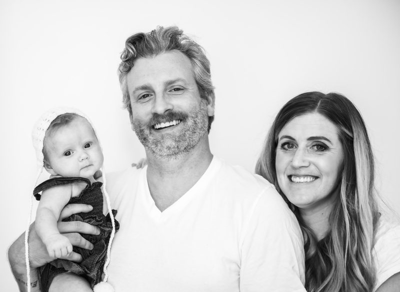 Family of three portrait against a white backdrop, newborn in a knit bonnet, everyone looking at the camera