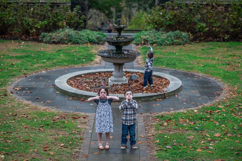Two kids making silly faces by a garden fountain while their little brother points at the sky, photographed candidly in a Philadelphia park