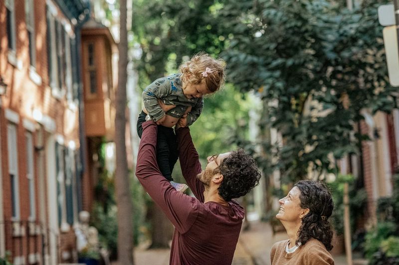Father lifting his toddler overhead on a Philadelphia street while mom laughs — a candid moment from a family photo session