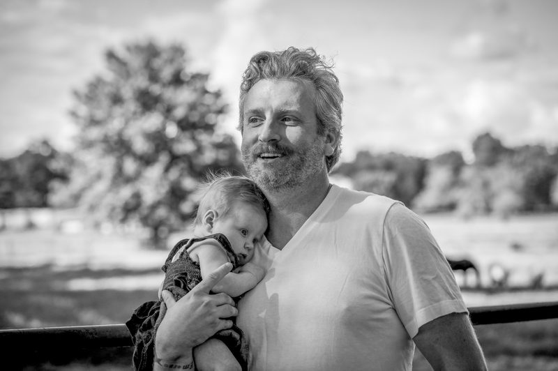 Black-and-white portrait of a father smiling with his newborn tucked under his chin at a horse farm