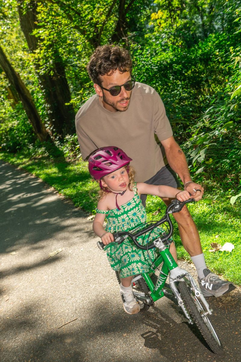 Father jogging alongside his daughter as she pedals a small green bike down a shaded path