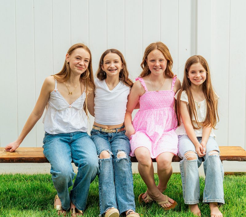 Four sisters in white tops and jeans seated along a wooden bench in summer