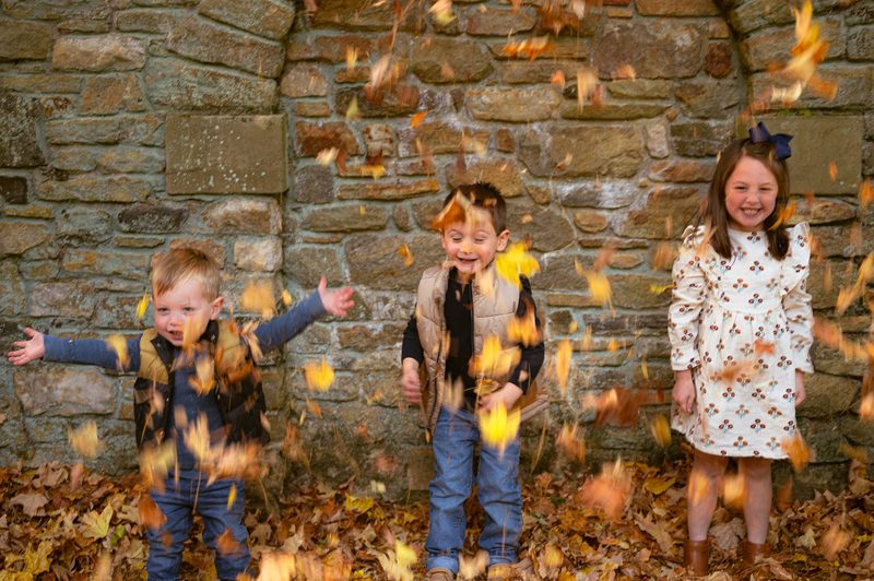 Three children flinging armfuls of autumn leaves in front of a stone wall during a Philadelphia family session