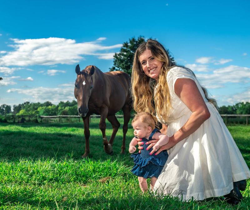 Mother and baby in a sunny pasture with a chestnut horse walking up behind them during a family session
