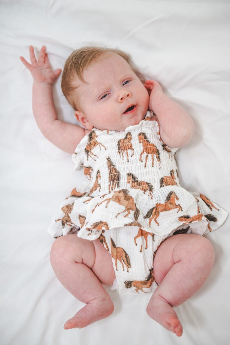 Newborn stretching one arm overhead on a white sheet in a horse-print romper