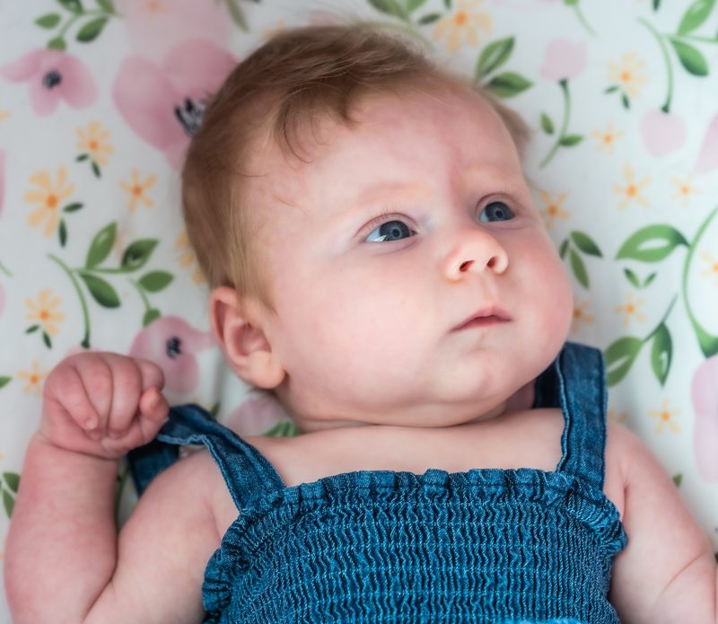 Close-up of a newborn in a denim romper lying on a floral blanket, gazing off to the side