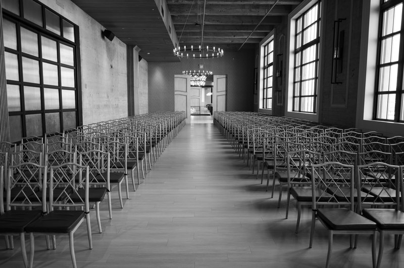 Empty wedding ceremony hall in black and white, rows of chairs receding toward tall windows under a concrete ceiling