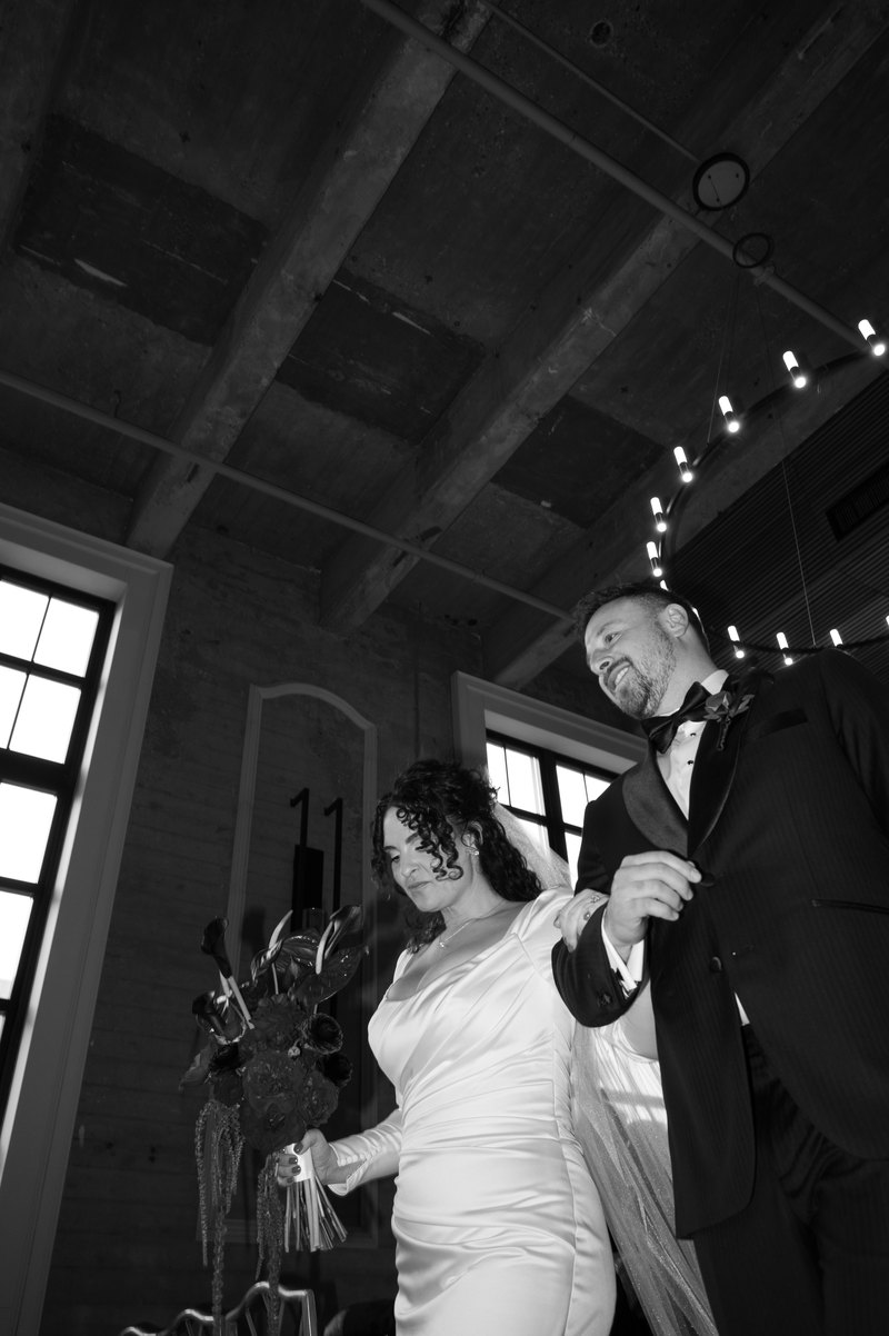 Bride and groom moving together on the dance floor beneath a ring of string lights in a converted industrial hall