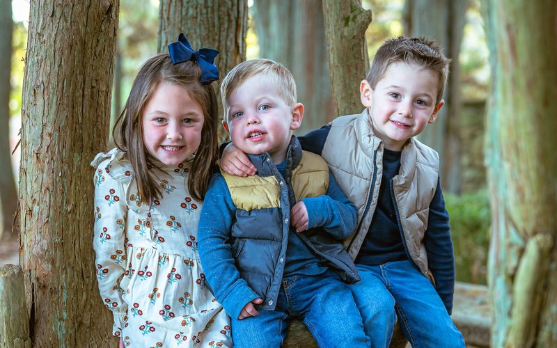 Three young siblings sitting together at the base of a tree in autumn woods near Philadelphia