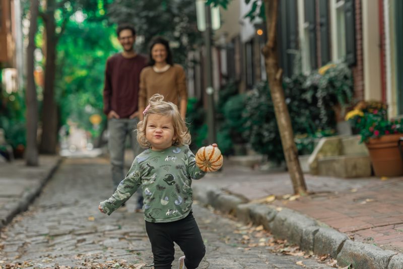 Toddler carrying a basketball down a cobblestone Philadelphia street while her parents follow behind