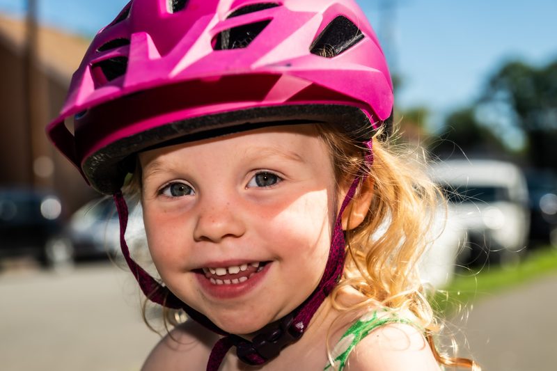 Close-up of a toddler laughing in a pink bike helmet, curls lit by low sun