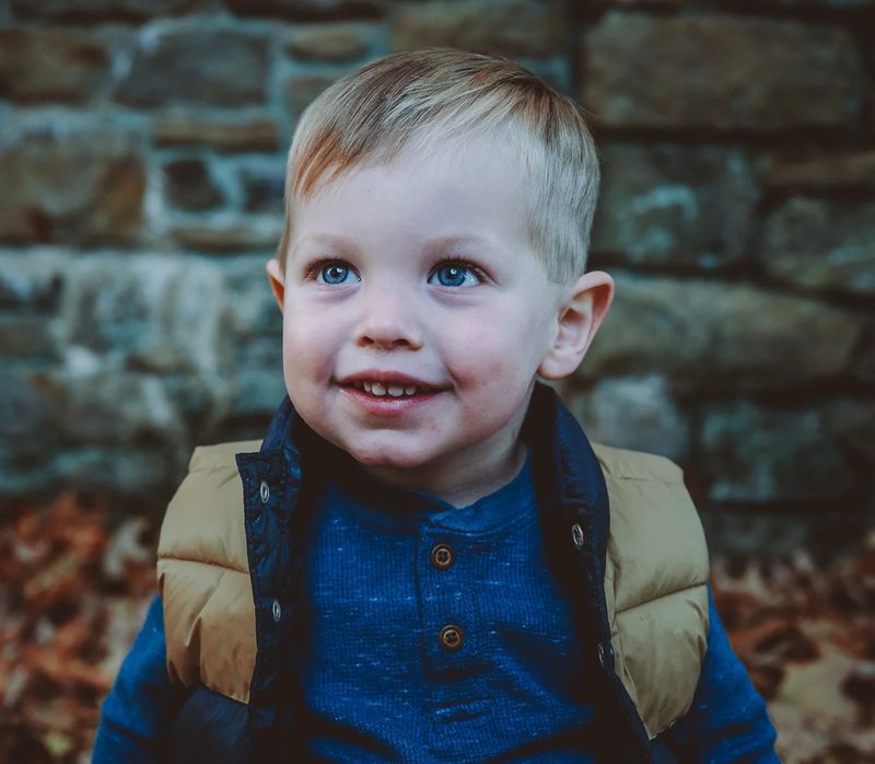 Close-up portrait of a blue-eyed toddler in a tan puffy vest smiling in front of a stone wall